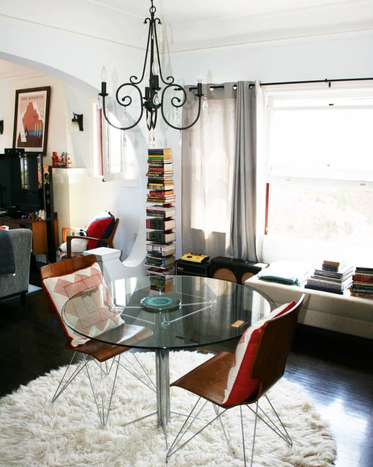 Glass dining table with wooden chairs on a fluffy rug, under a black chandelier, surrounded by books and gray curtains.