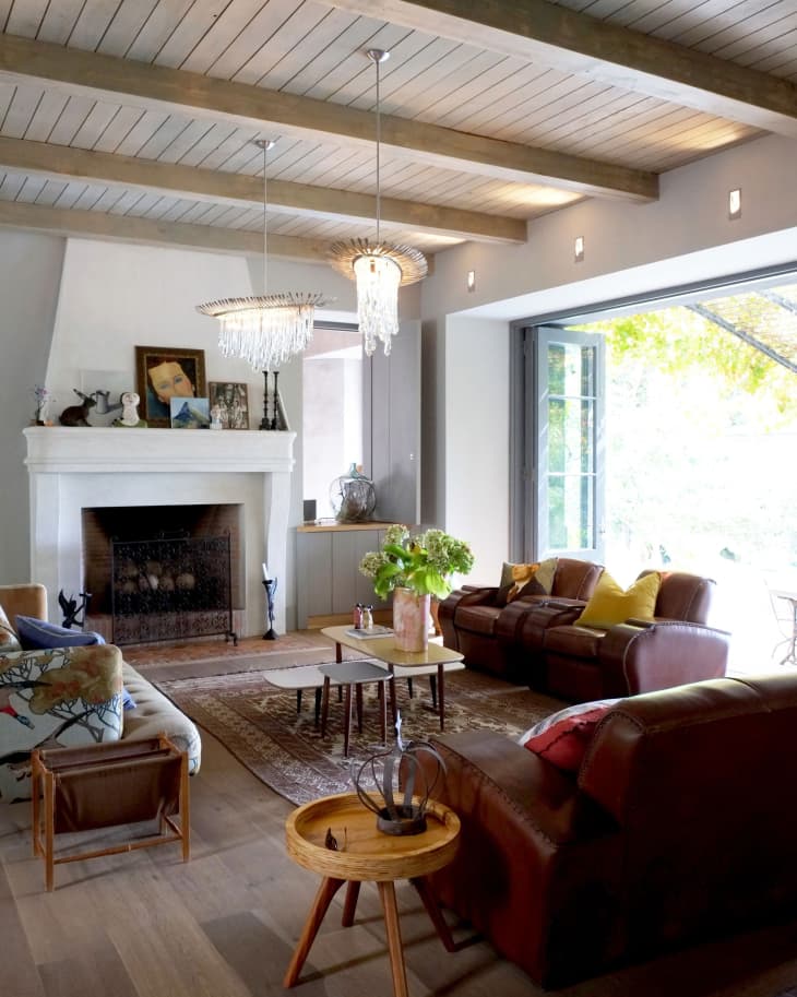 Living room with brown leather sofas, wooden coffee table, fireplace, and chandelier, featuring large windows and green plants.