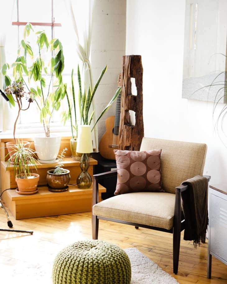 Cozy living room with a beige armchair, green knitted pouf, potted plants, and a guitar by the window.