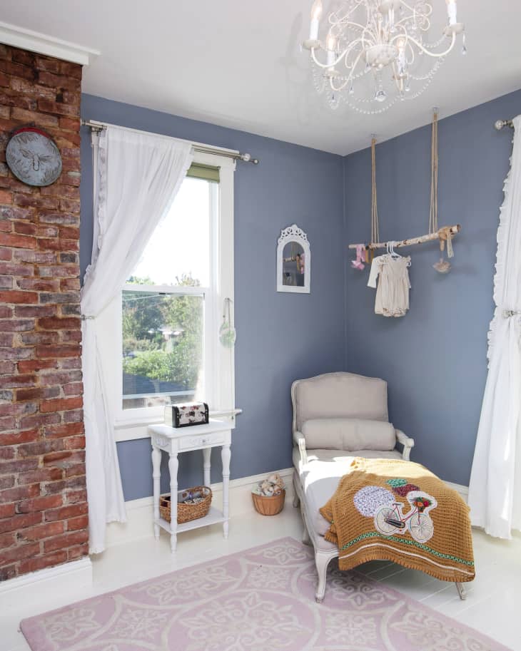 Nursery with blue walls, white armchair, chandelier, and a decorative blanket on a pink patterned rug.