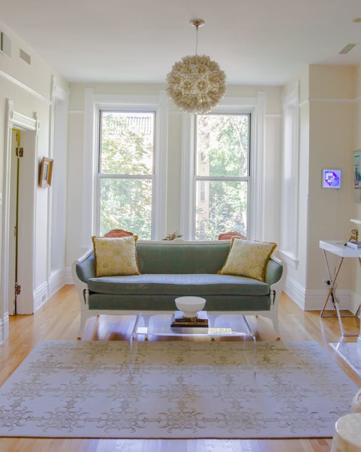 Living room with a green sofa, yellow pillows, floral chandelier, and large windows overlooking trees.