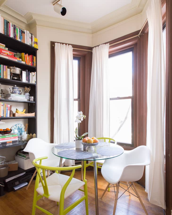 Cozy dining nook with round glass table, white and green chairs, bookshelves, and sheer curtains.