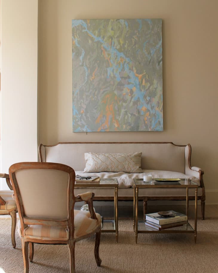 Elegant living room with a beige sofa, abstract painting, glass coffee table, and upholstered chairs on a woven rug.