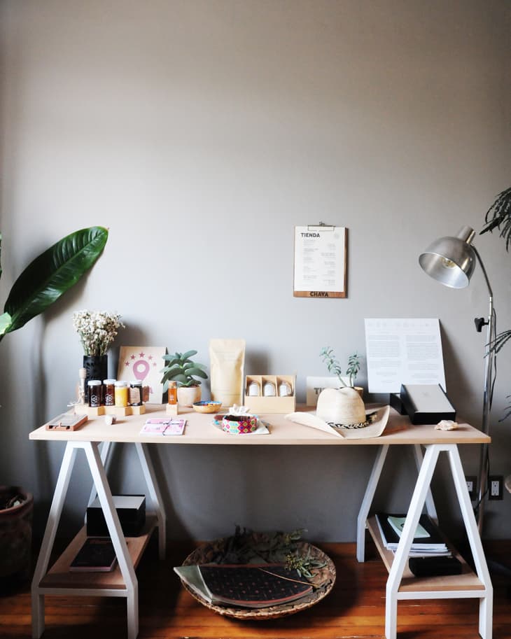 Wooden desk with assorted items, including plants, jars, a hat, and a lamp, against a gray wall with a menu.