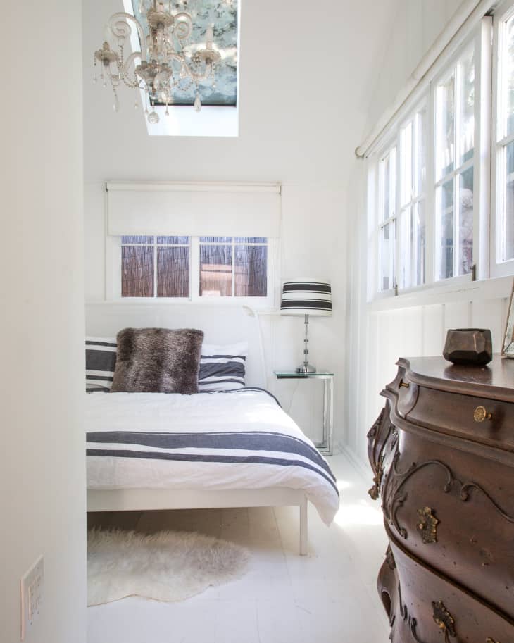 Small bedroom with a skylight, chandelier, striped bedding, fur pillows, and a vintage wooden dresser.