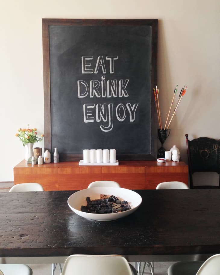 Chalkboard with "Eat Drink Enjoy" above a wooden sideboard, vase of flowers, and a bowl on a dark dining table.