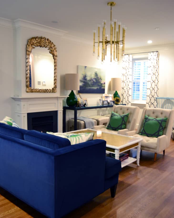 Living room with blue sofa, beige armchairs, green pillows, gold chandelier, and decorative mirror above fireplace.