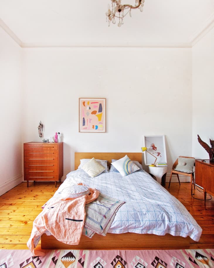 Bedroom with wooden bed, colorful pillows, abstract wall art, pink patterned rug, and vintage chandelier.