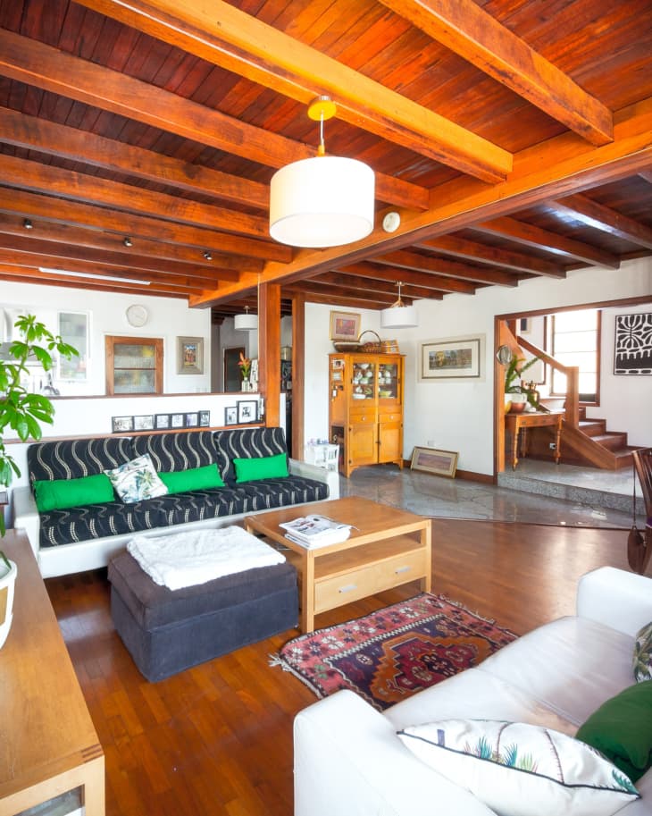 Cozy living room with wooden ceiling, patterned sofa, green cushions, and a colorful rug, leading to a staircase.