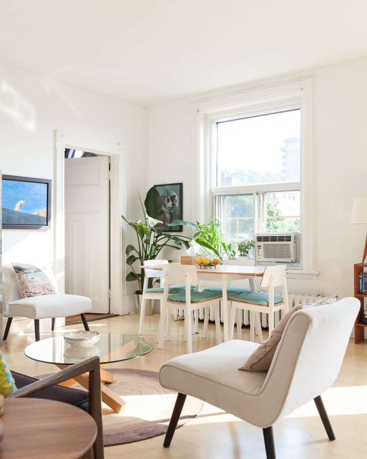 Bright living room with white chairs, glass coffee table, dining set, large window, and green plants.