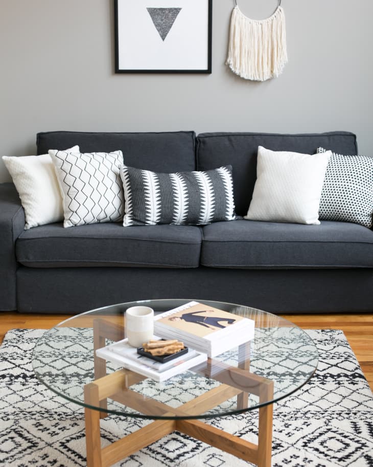 Gray sofa with patterned cushions, glass coffee table, and geometric rug in a modern living room with wall art and macrame.