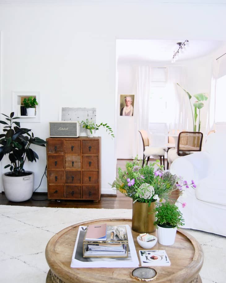 Living room with round wooden coffee table, books, plants, and a vintage cabinet. Dining area visible in the background.