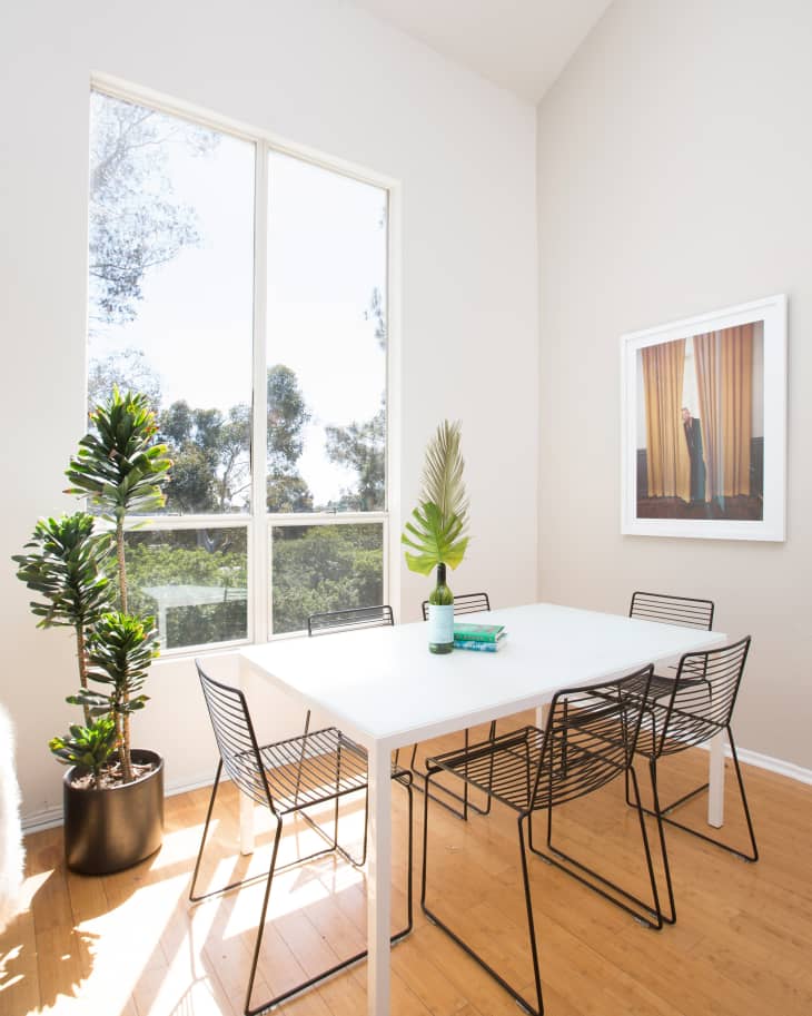 Bright dining room with a white table, black wire chairs, large window, potted plants, and framed wall art.