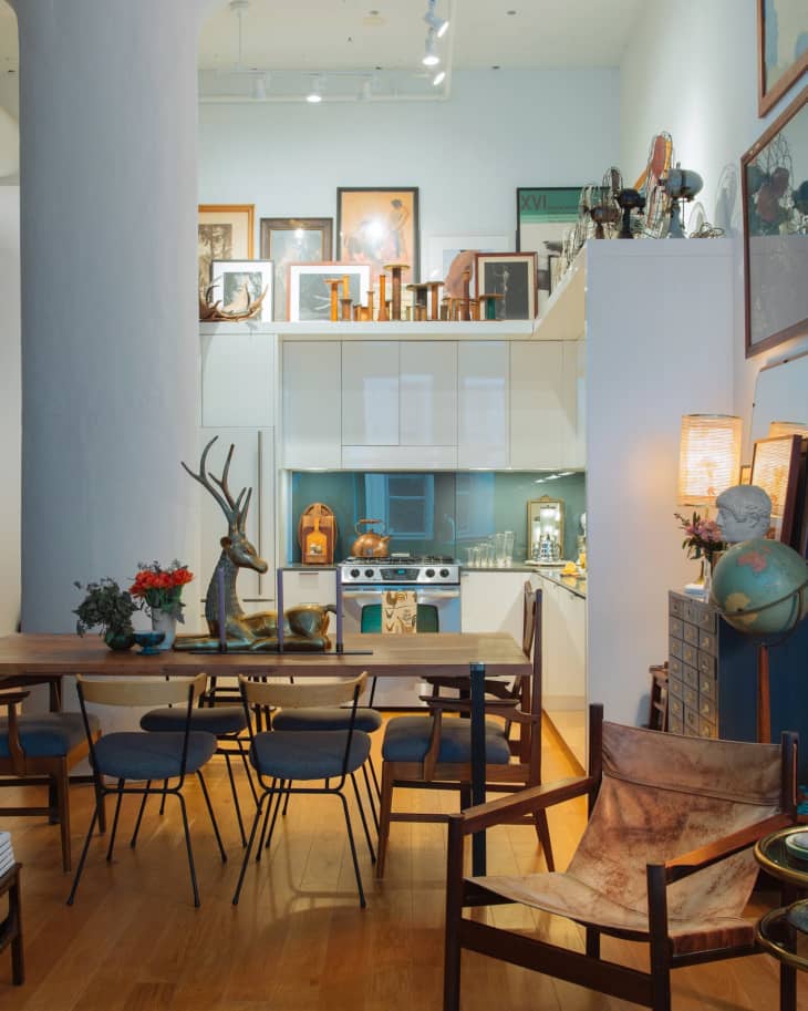 Open-plan kitchen and dining area with wooden table, blue chairs, eclectic decor, and framed art on white cabinets.