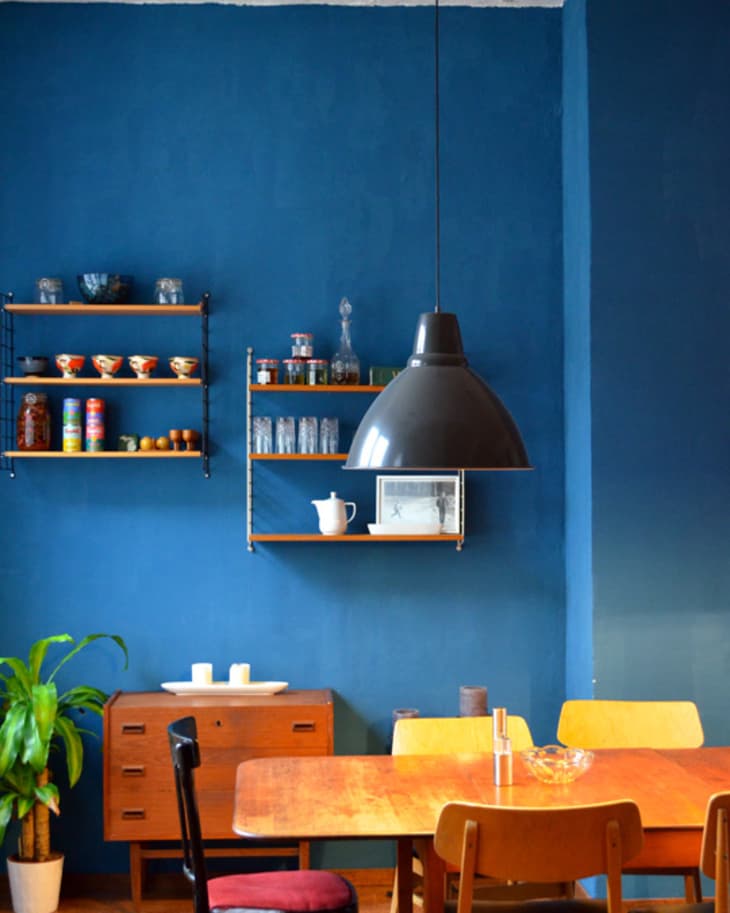 Dining area with wooden table, mixed chairs, blue wall, shelves with dishes, and a large black pendant light.