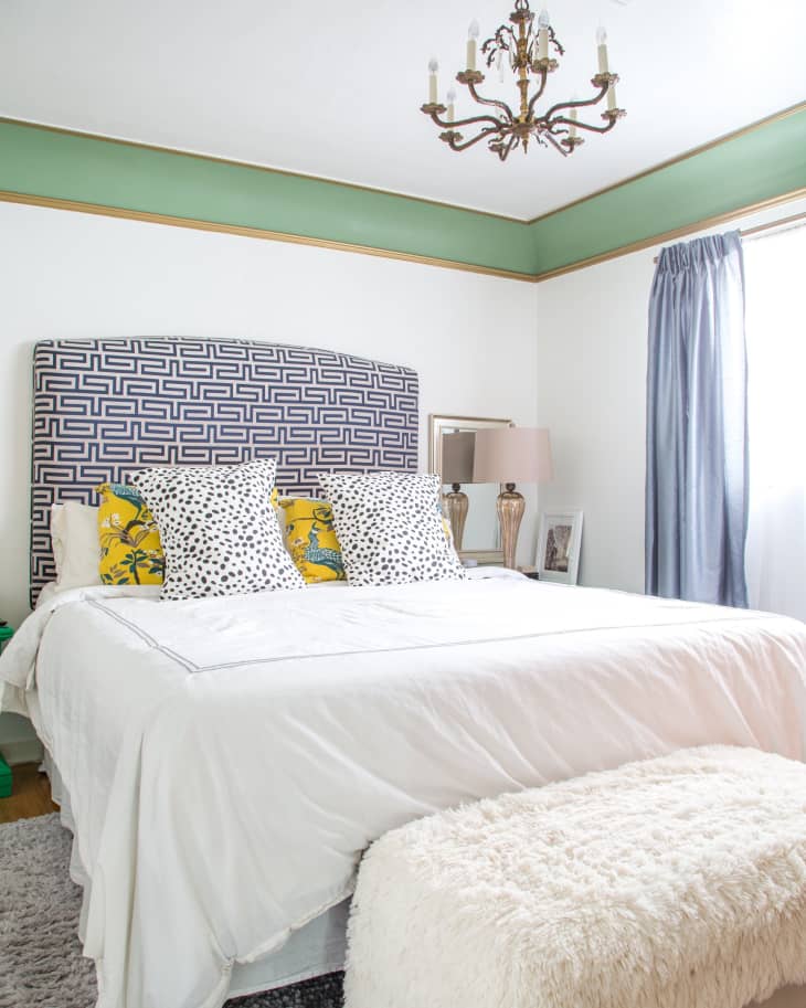 Bedroom with a patterned headboard, white bedding, yellow and polka dot pillows, chandelier, and a fluffy bench.