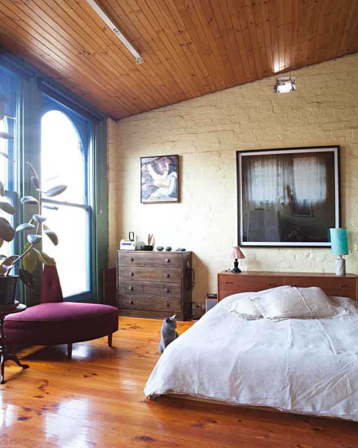 Bedroom with wooden ceiling, large window, red chair, wooden dresser, bed with white linens, and a cat on the floor.