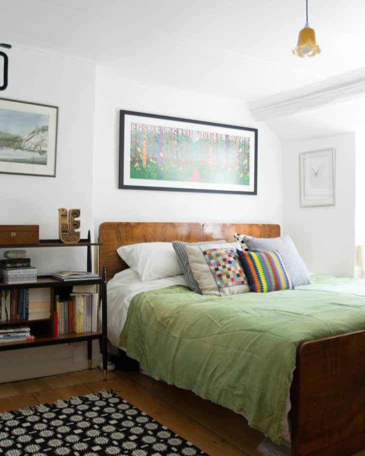 Bedroom with wooden bed, green bedding, colorful pillows, bookshelf, and framed artwork on white walls.