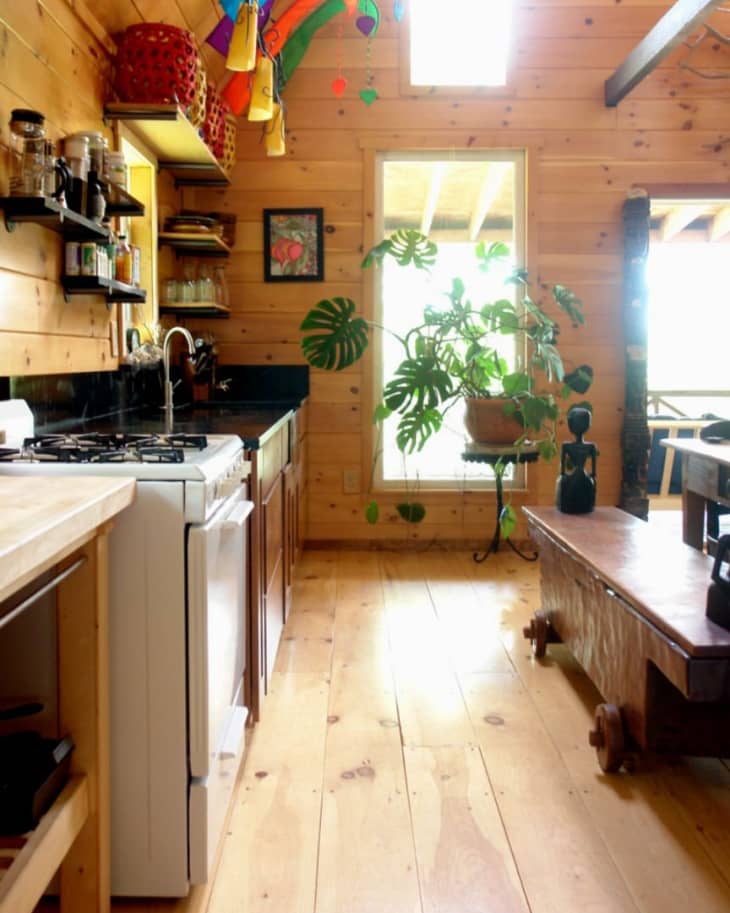 Wooden kitchen with open shelves, colorful hanging decor, potted plant, and a rustic bench under a skylight.