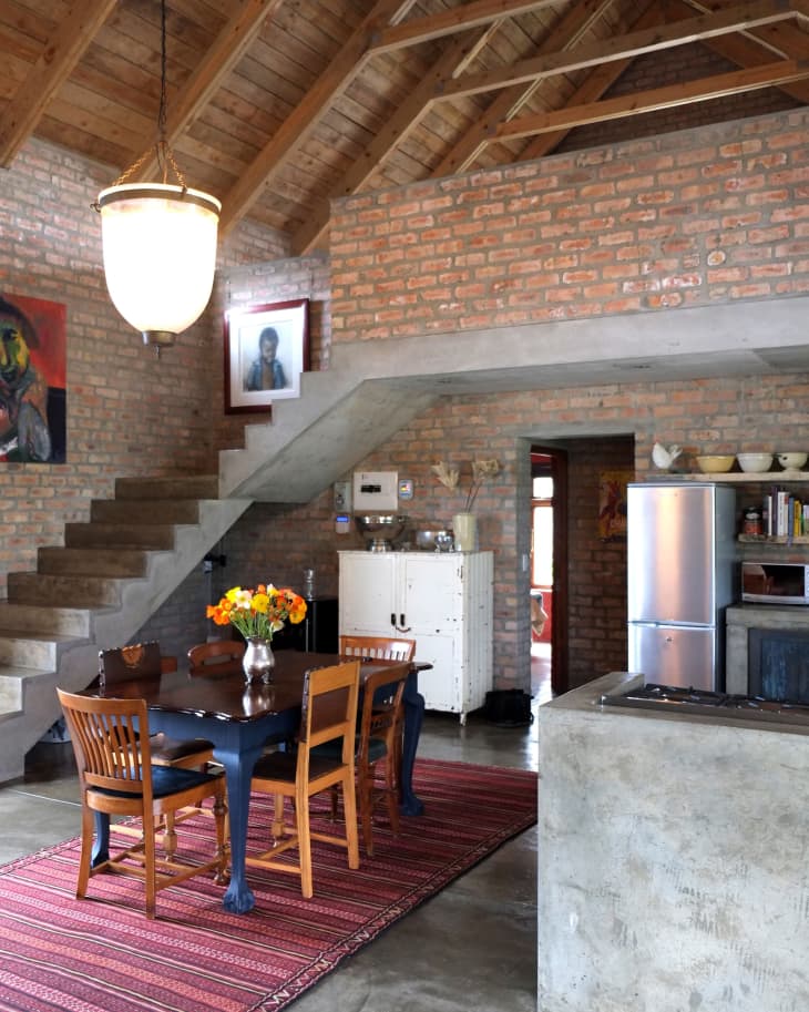 Rustic dining area with wooden table, chairs, brick walls, exposed beams, and a red patterned rug.