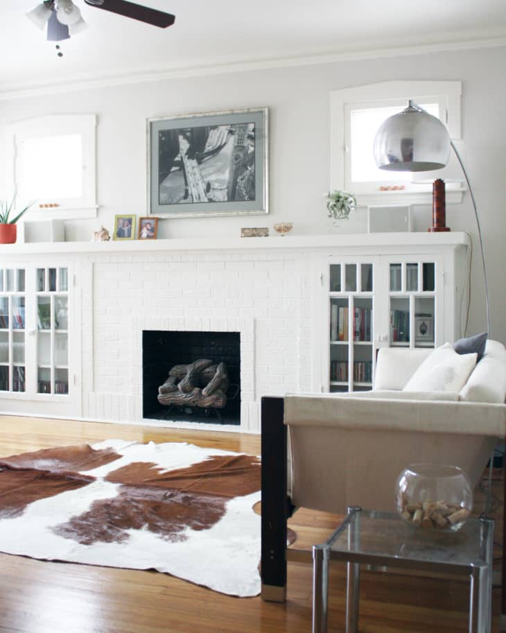 Living room with white brick fireplace, cowhide rug, beige sofa, glass side table, and ceiling fan.