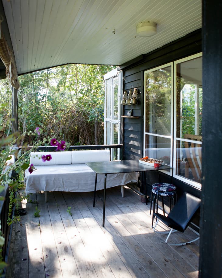 Covered porch with a white bench, black table, floral stool, and potted plants, surrounded by greenery.