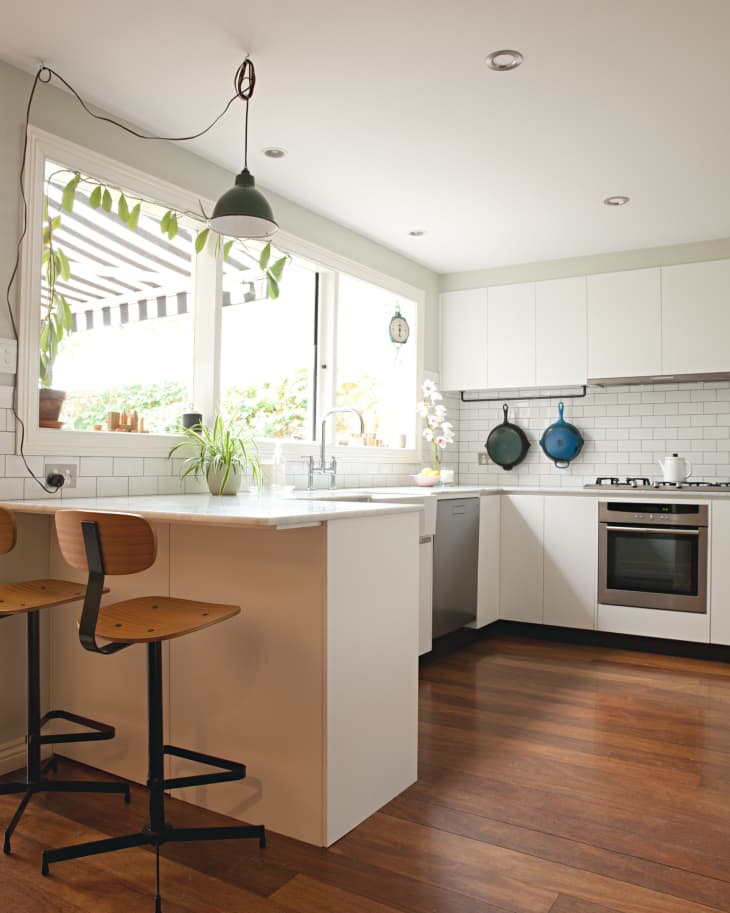 Modern kitchen with white cabinets, wooden stools, hanging plants, and a large window with striped awning.