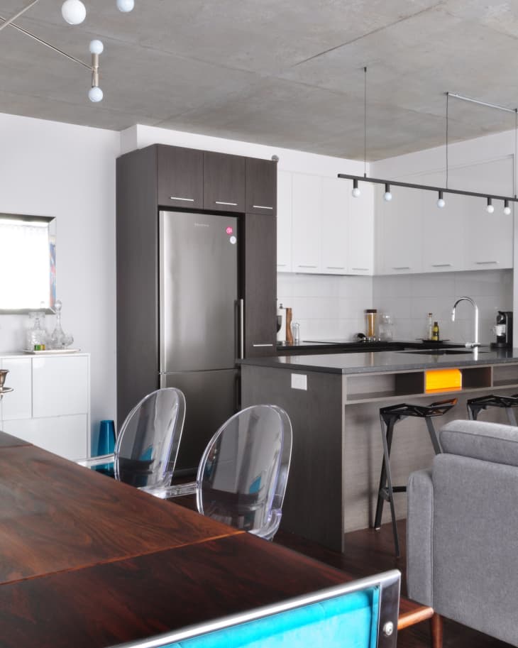 Modern kitchen with dark cabinets, stainless steel fridge, island with bar stools, and clear dining chairs.