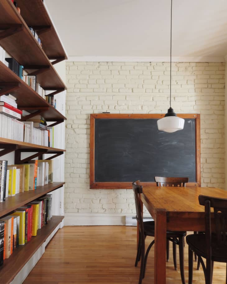 Dining room with wooden table, chairs, brick wall, chalkboard, and bookshelves.