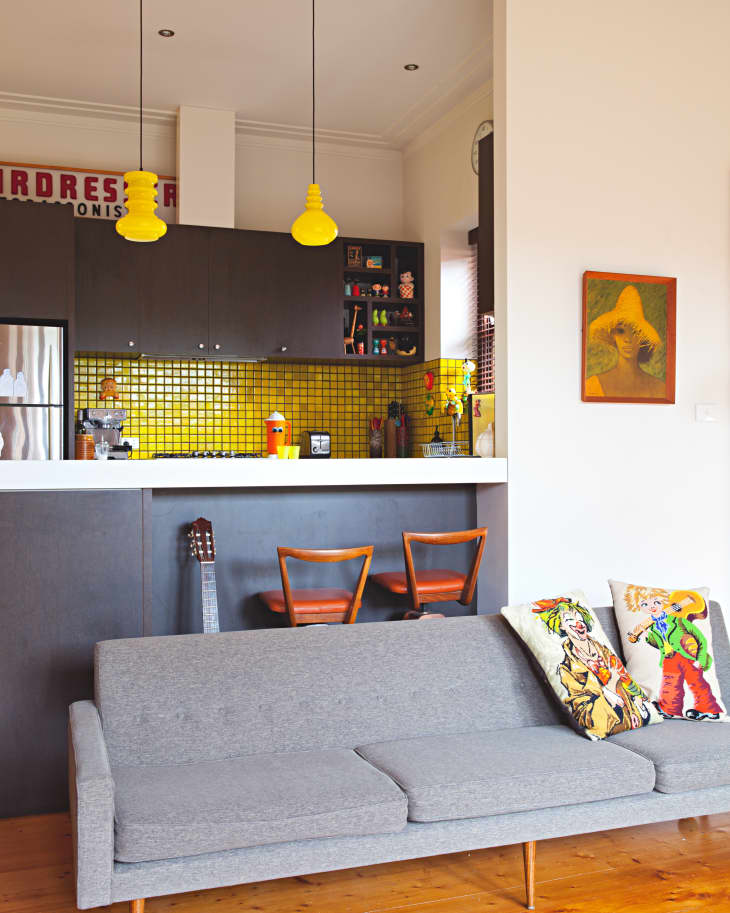 Gray sofa with colorful pillows, wooden floor, yellow-tiled kitchen, and pendant lights in a modern living space.