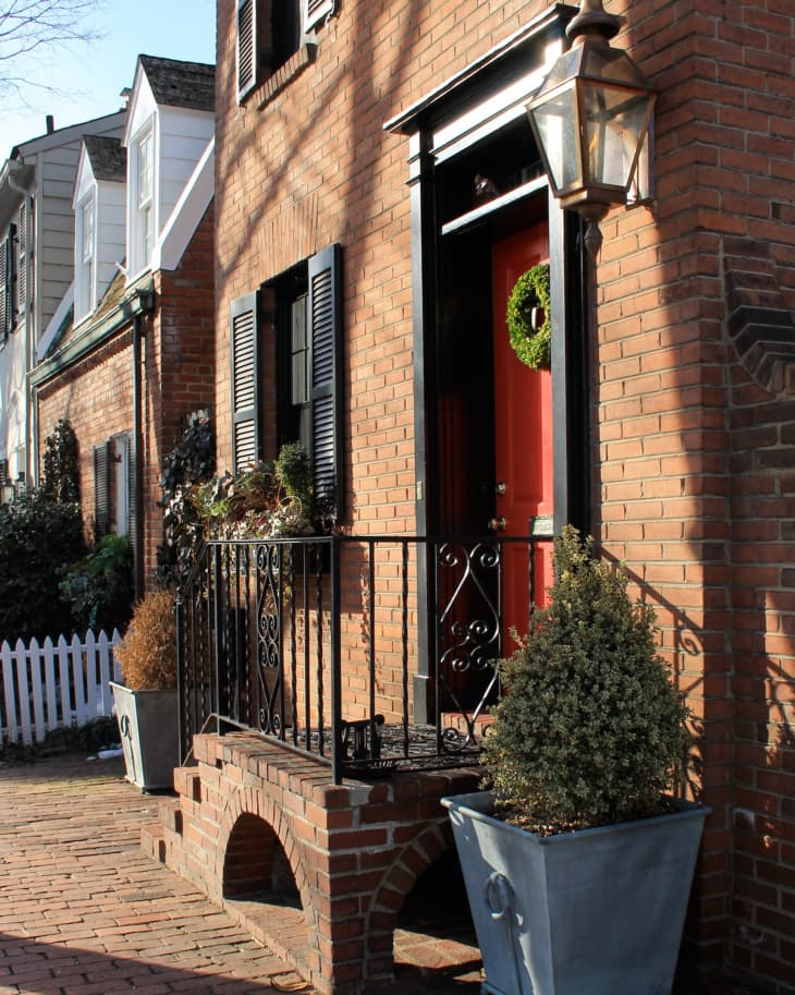 Brick townhouse with red door, black shutters, wrought iron railing, and potted plants on a brick walkway.