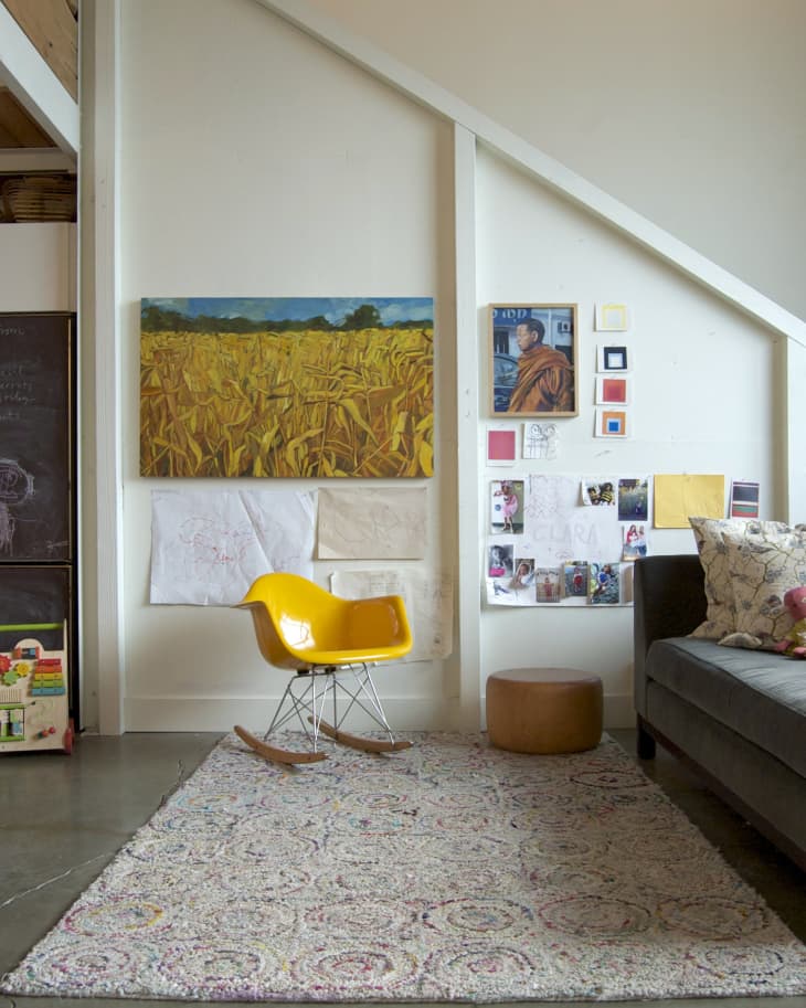 Living room with yellow rocking chair, abstract wall art, gray sofa, and patterned rug on concrete floor.