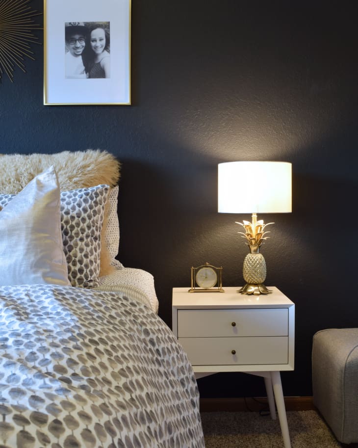 Bedroom with dark wall, white nightstand, pineapple lamp, framed photo, and patterned bedding.