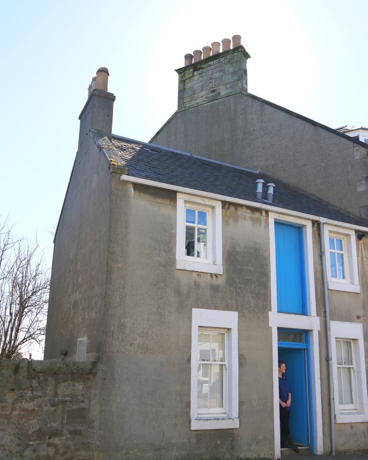 Stone house with blue door and windows, person standing in doorway, chimney pots on roof.