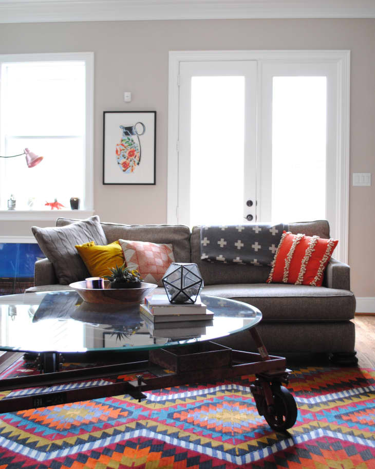 Living room with gray sofa, colorful pillows, glass coffee table, geometric decor, and vibrant patterned rug.