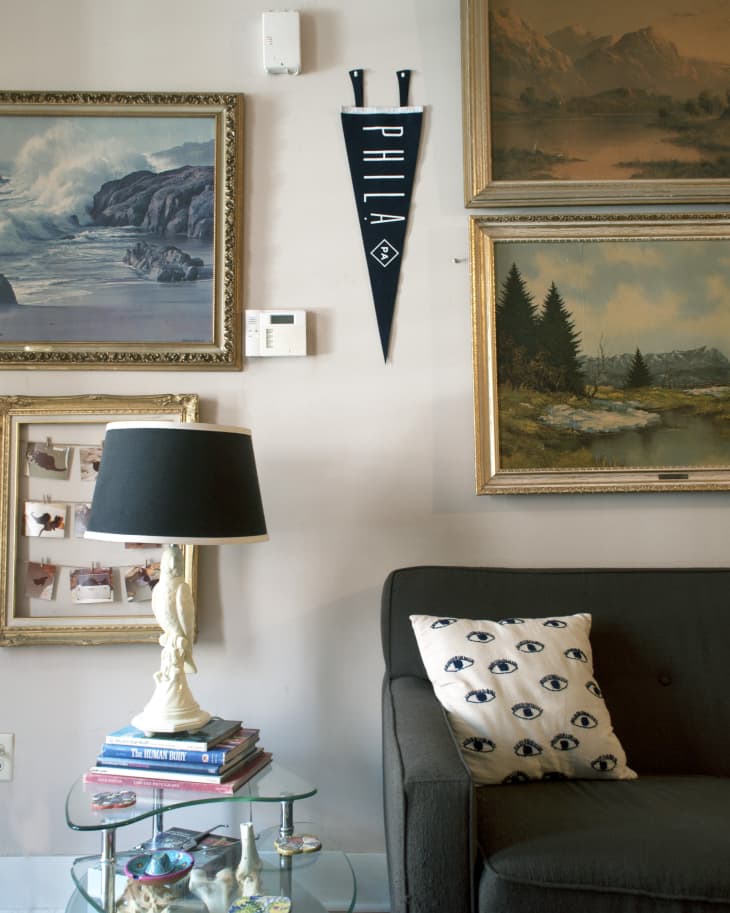 Living room with framed paintings, a black pennant, glass table, books, and a gray sofa with an eye-patterned pillow.