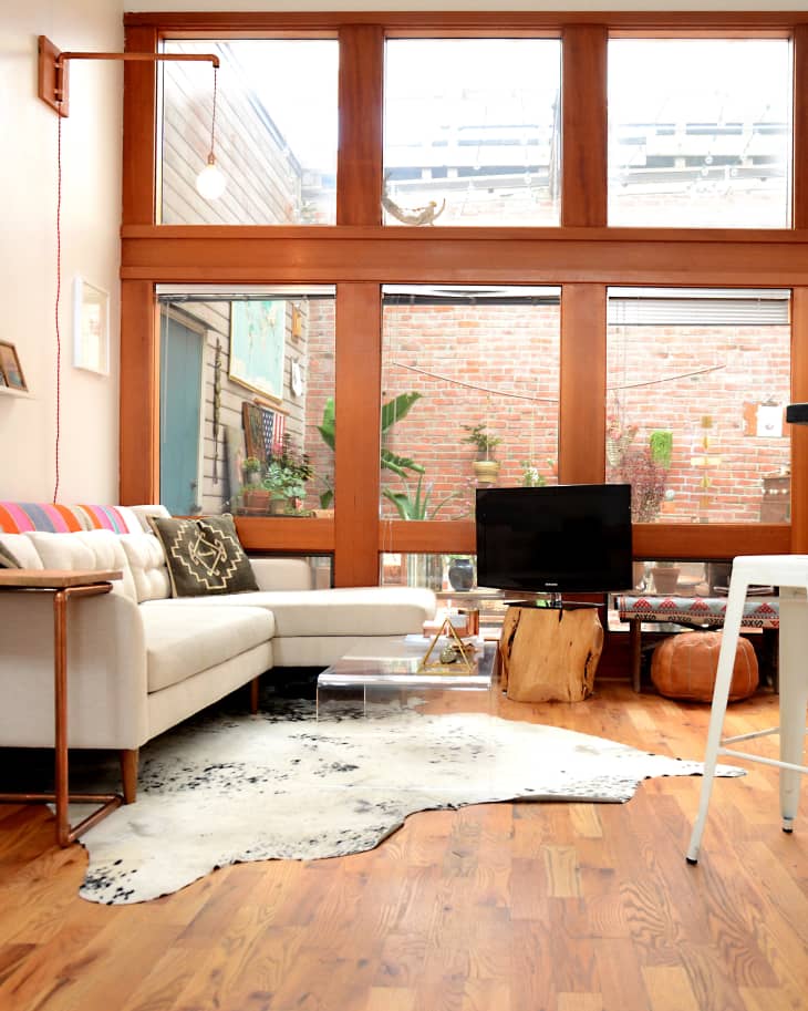 Living room with a cream sofa, colorful cushions, cowhide rug, and large windows overlooking a brick wall and plants.