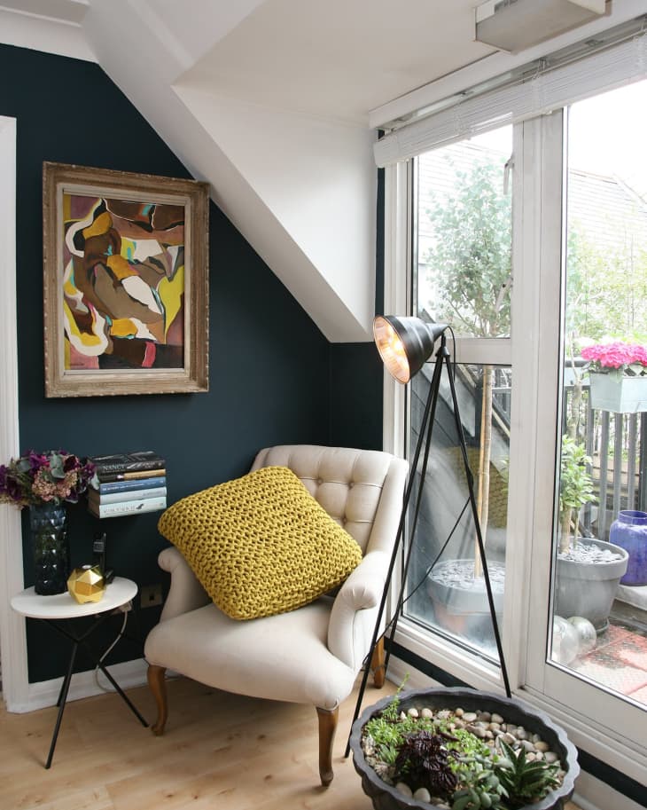 Cozy reading nook with a beige armchair, yellow cushion, floor lamp, abstract painting, and potted plants by a window.