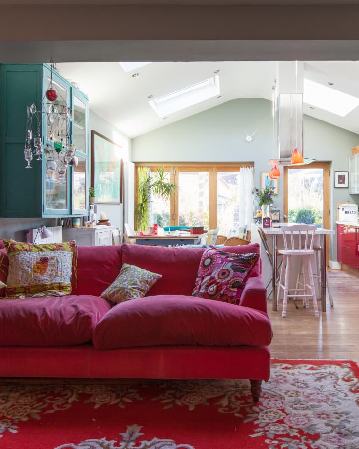 Red sofa with patterned cushions in a colorful living room, featuring a red rug, green cabinets, and a bright dining area.
