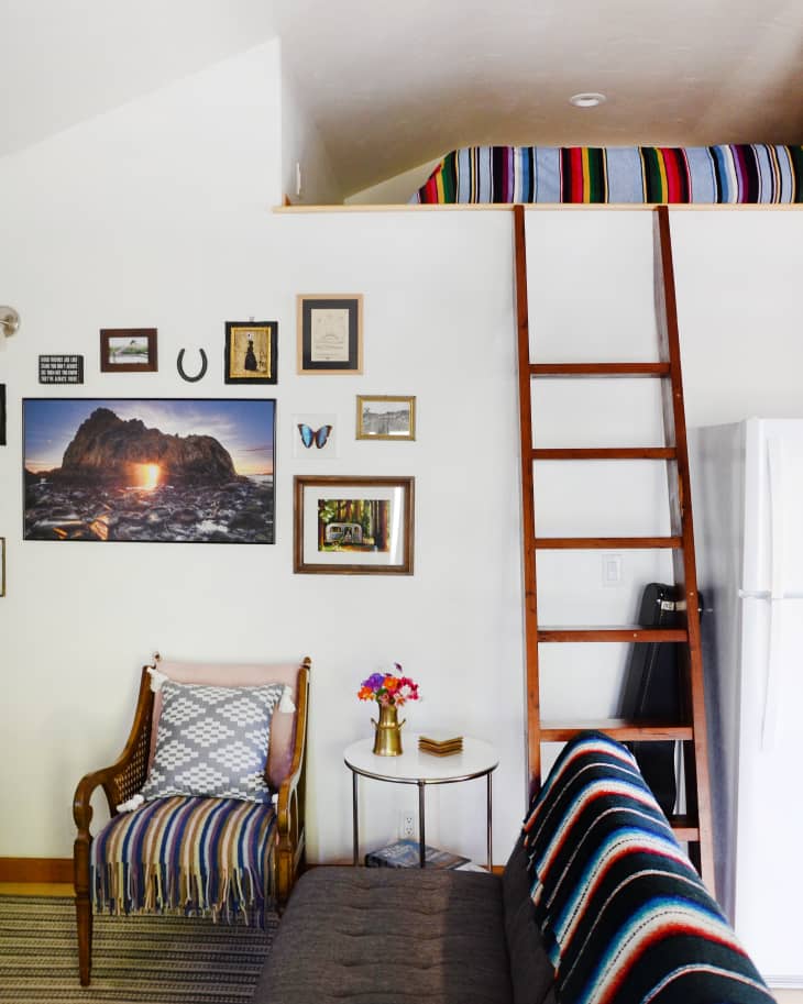 Living room with a loft bed, wooden ladder, framed wall art, striped blankets, and a small table with flowers.