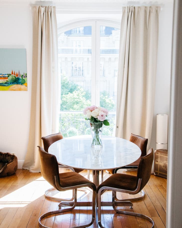 Round marble dining table with brown chairs, vase of pink flowers, and large window with curtains in a bright room.