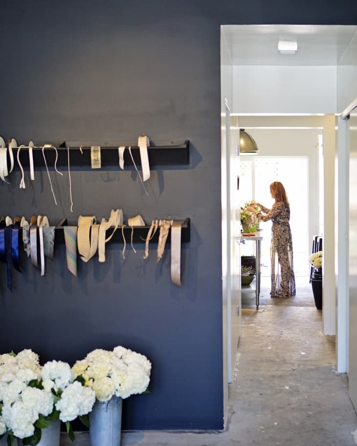 Dark wall with hanging ribbons, metal buckets of white flowers, and a person arranging flowers in a bright room.