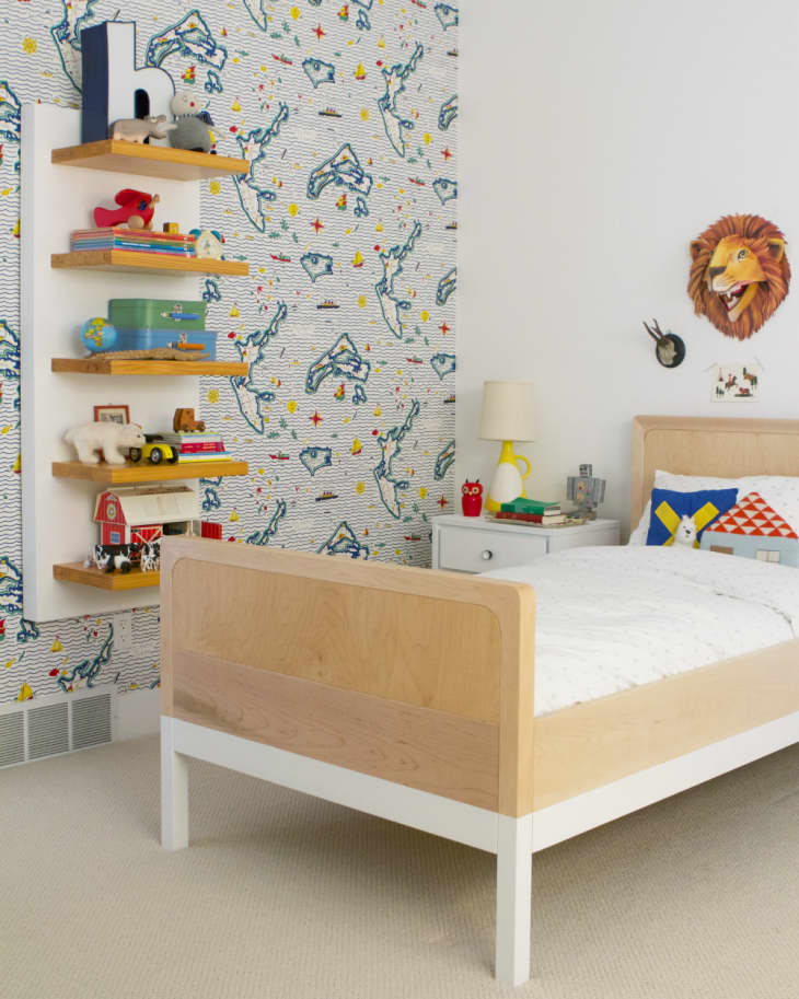 Child's bedroom with a wooden bed, nautical-themed wallpaper, floating shelves with toys, and a lion wall decoration.