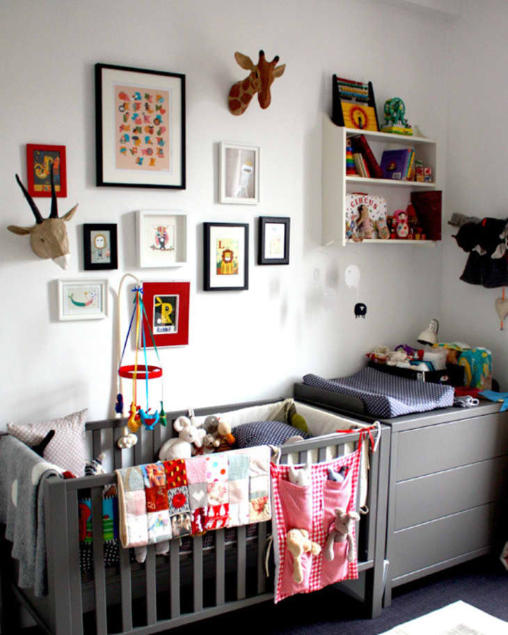 Gray crib with colorful toys and blankets, wall art, and shelves with books and stuffed animals in a nursery.