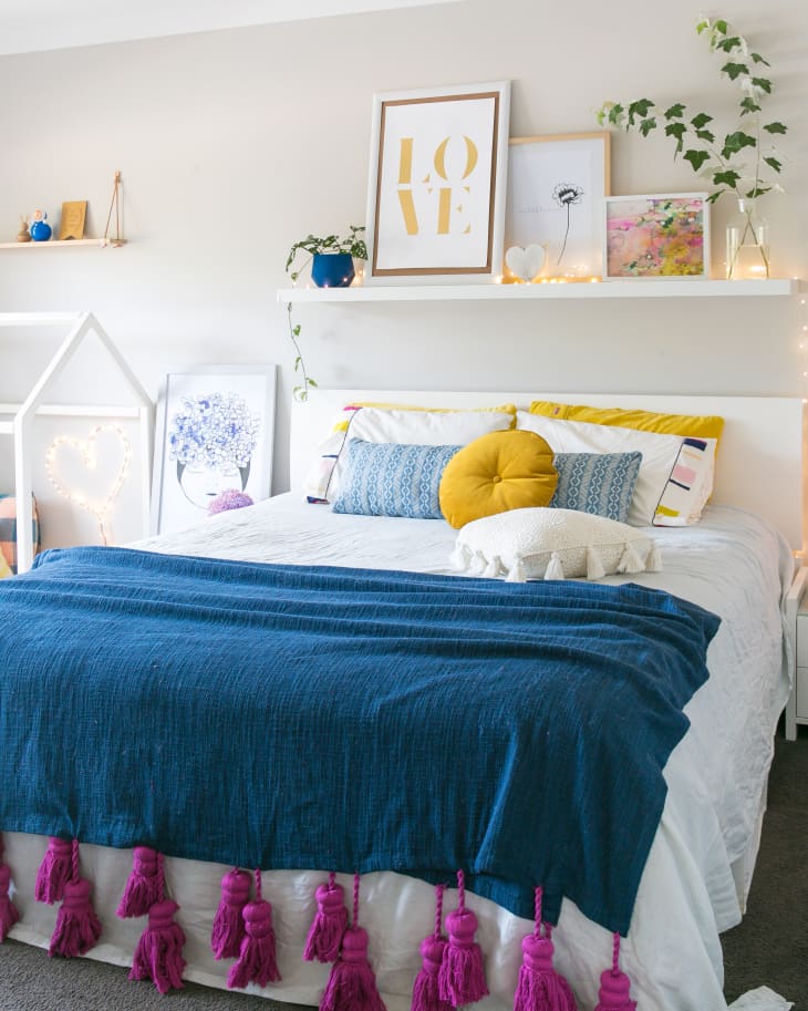 Cozy bedroom with a blue throw, pink tassels, yellow pillow, and decorative shelf with framed art and plants.