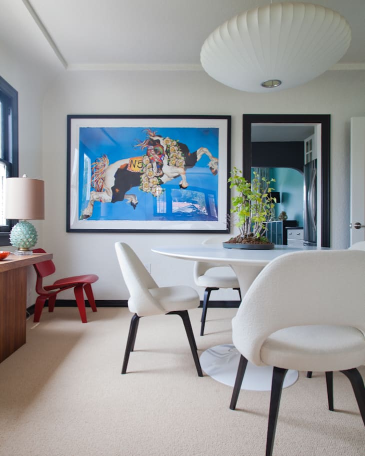 Dining room with white chairs, round table, large artwork, red stool, and pendant light.