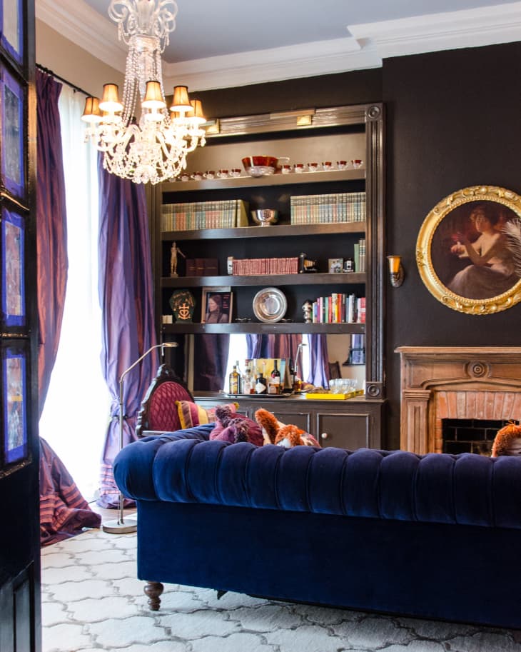 Elegant living room with a blue velvet sofa, chandelier, bookshelves, and a fireplace adorned with a portrait.