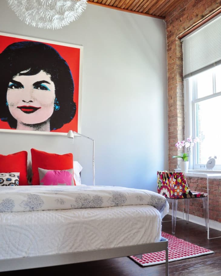 Bedroom with pop art portrait, red pillows, floral chair, and modern chandelier.