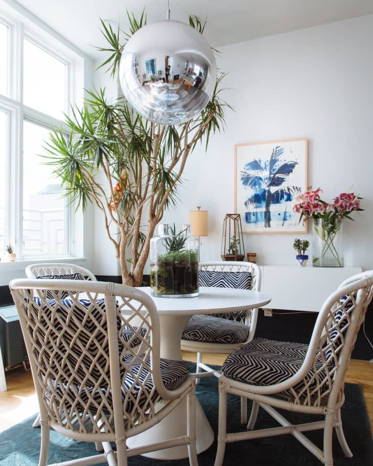 Dining area with white wicker chairs, round table, large plant, silver pendant light, and abstract blue artwork.