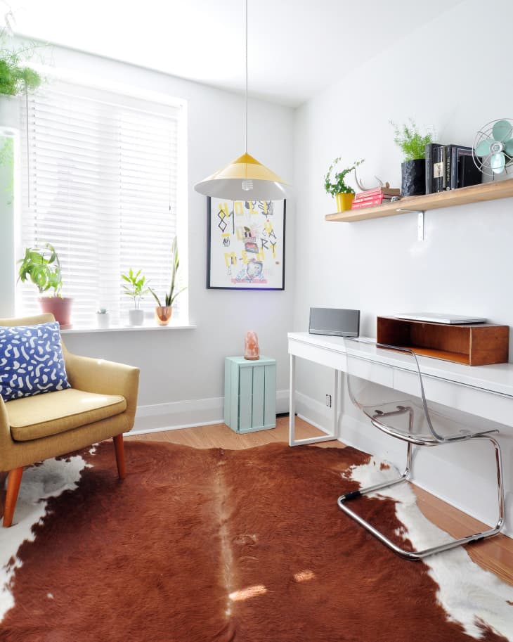 Home office with a yellow chair, brown cowhide rug, white desk, clear chair, and plants on a shelf and windowsill.